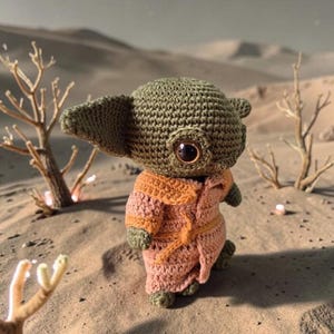 Op de afbeelding: Een gehaakte figuur van een personage uit een populaire sciencefiction-serie. De figuur is voornamelijk olijfgroen met grote, donkere ogen en draagt een roze en oranje gewaad. De achtergrond toont een woestijnlandschap met schaarse vegetatie.