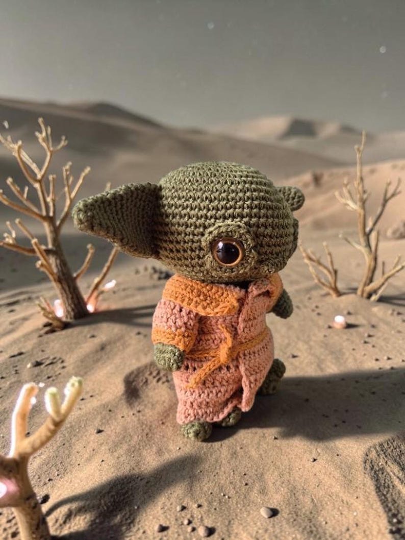 Op de afbeelding: Een gehaakte figuur van een personage uit een populaire sciencefiction-serie. De figuur is voornamelijk olijfgroen met grote, donkere ogen en draagt een roze en oranje gewaad. De achtergrond toont een woestijnlandschap met schaarse vegetatie.