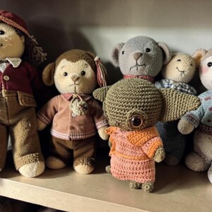 Op de afbeelding: Een verzameling handgemaakte gehaakte poppen, waaronder een aap, een beer en een uniek wezen met grote ogen. De poppen zijn in verschillende kleuren, met gedetailleerde kleding en accessoires, tentoongesteld op een houten plank.