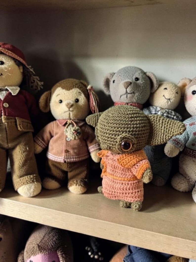 Op de afbeelding: Een verzameling handgemaakte gehaakte poppen, waaronder een aap, een beer en een uniek wezen met grote ogen. De poppen zijn in verschillende kleuren, met gedetailleerde kleding en accessoires, tentoongesteld op een houten plank.