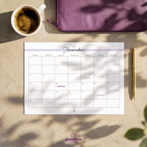 May include: A 2026 November calendar page with a white background, black grid lines, and a light purple header. The text includes the month name, days of the week in German, and dates. A gold pen is positioned to the right of the calendar sheet.