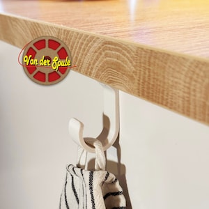 Op de afbeelding: Een witte plastic haak gemonteerd onder een houten kast met een gestreepte keukendoek er aan hangend.