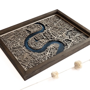 Puede incluir: Una pieza de arte de pared enmarcada en madera con un mapa 3D de una ciudad cortado con láser, con un río que serpentea a través de ella. El mapa está en tonos marrones y azules, y el marco es de madera oscura.