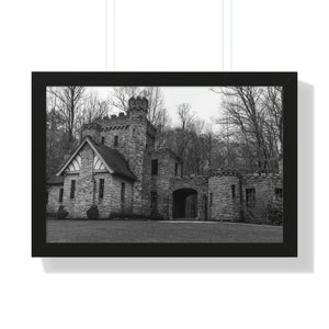 May include: Black and white framed print of a stone castle with turrets and arched entryways. The castle is surrounded by trees and set against a cloudy sky. The print is in a black frame.