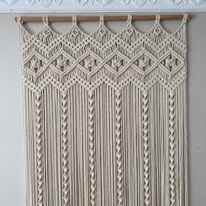 May include: Cream-colored macrame wall hanging with a geometric design. The top features a series of diamond shapes, and the bottom has long, vertical strands. The macrame is suspended from a wooden rod, adding a natural touch to the decor.