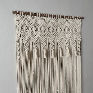 May include: Cream-colored macrame wall hanging. The top features a geometric pattern with diamond shapes and tassels. Vertical strands of knotted rope hang below, creating a textured, bohemian-style decor piece. The macrame is mounted on a wooden rod.