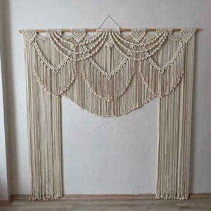 May include: A large, white macrame wall hanging with a wooden dowel at the top. The macrame is made of a series of intricate knots and patterns, creating a cascading effect. The hanging is perfect for adding a bohemian touch to any room.