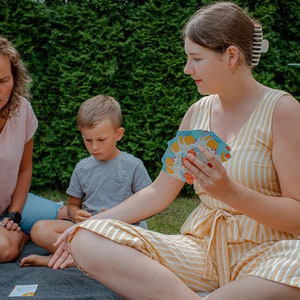 Op de afbeelding: Drie personen, twee volwassenen en een kind, zitten op een grijze deken buiten. De volwassenen spelen een kaartspel met kleurrijke kaarten. Het kind kijkt naar het spel. De volwassenen dragen casual kleding. Het kind draagt een grijs shirt. De achtergrond is een groene haag.