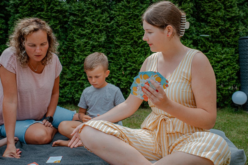 Op de afbeelding: Drie personen, twee volwassenen en een kind, zitten op een grijze deken buiten. De volwassenen spelen een kaartspel met kleurrijke kaarten. Het kind kijkt naar het spel. De volwassenen dragen casual kleding. Het kind draagt een grijs shirt. De achtergrond is een groene haag.
