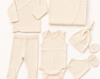 May include: A set of cream-colored baby clothing and accessories including a long-sleeve wrap top, footed pants, a sleeveless bodysuit, a folded blanket, a knotted beanie, a triangular bib, and a pair of small mittens, all in a solid neutral tone.