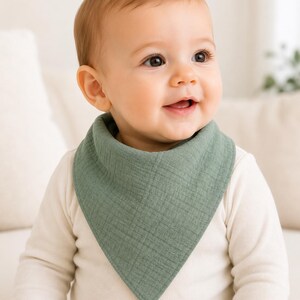 Puede incluir: Un babero triangular estilo bandana de color verde salvia, confeccionado en tela de muselina texturizada. El babero presenta un diseño sencillo de color sólido con costuras visibles en los bordes, usado alrededor del cuello sobre una camisa.