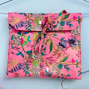 Pu&ograve; includere: Un completo pigiama rosa a fantasia floreale, piegato e legato con un nastro di tessuto abbinato. Il completo presenta un design vibrante di foglie verdi, fiori blu e bianchi e accenti rosa. Il completo &egrave; appeso a una gruccia argentata su uno sfondo azzurro.