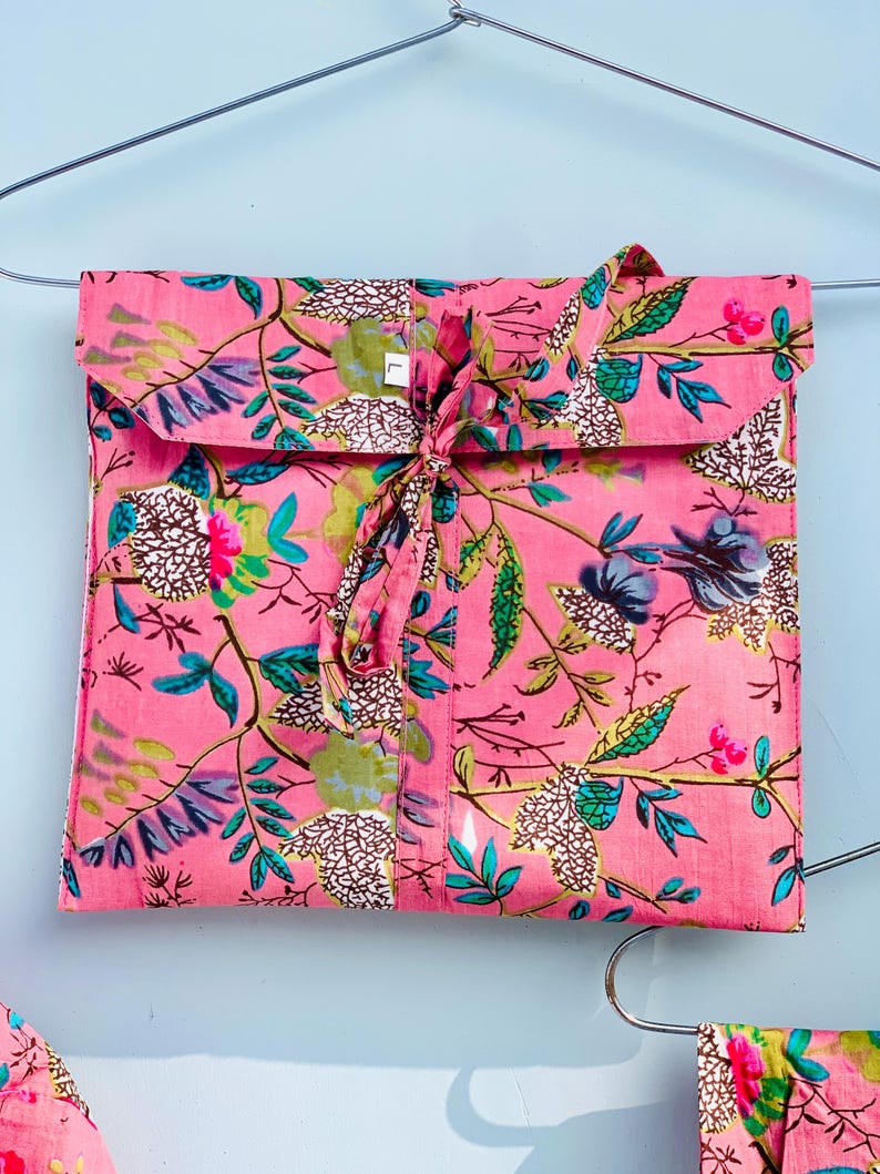 Pu&ograve; includere: Un completo pigiama rosa a fantasia floreale, piegato e legato con un nastro di tessuto abbinato. Il completo presenta un design vibrante di foglie verdi, fiori blu e bianchi e accenti rosa. Il completo &egrave; appeso a una gruccia argentata su uno sfondo azzurro.
