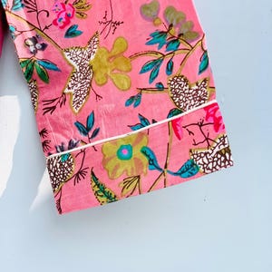 Pu&ograve; includere: Tessuto rosa con motivo floreale. I fiori sono gialli, rosa e blu, con foglie e steli verdi. Il tessuto ha un bordo bianco. Il design &egrave; un primo piano di un capo d'abbigliamento.