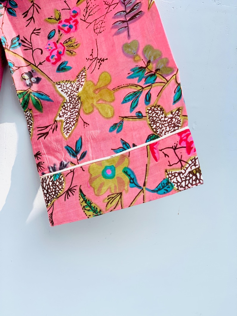 Pu&ograve; includere: Tessuto rosa con motivo floreale. I fiori sono gialli, rosa e blu, con foglie e steli verdi. Il tessuto ha un bordo bianco. Il design &egrave; un primo piano di un capo d'abbigliamento.