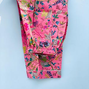 Pu&ograve; includere: Pantaloni pigiama rosa con motivo floreale. I pantaloni presentano un bordo bianco sull'orlo inferiore e una vita elasticizzata. Il design floreale include fiori e foglie rosa, blu, verdi e bianche.