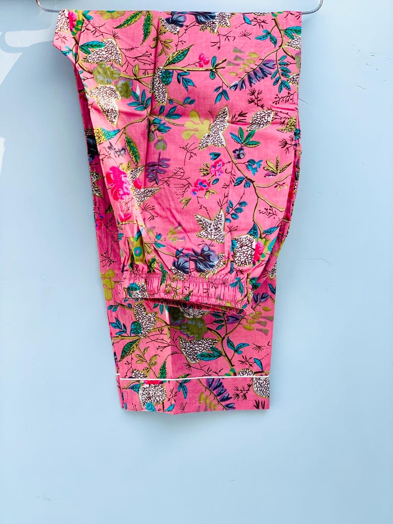 Pu&ograve; includere: Pantaloni pigiama rosa con motivo floreale. I pantaloni presentano un bordo bianco sull'orlo inferiore e una vita elasticizzata. Il design floreale include fiori e foglie rosa, blu, verdi e bianche.
