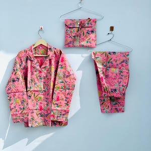 Pu&ograve; includere: Set pigiama rosa floreale, composto da una camicia con bottoni, pantaloni e una pochette abbinata. Il set presenta una stampa botanica con accenti verdi, gialli e bianchi. I pigiami sono esposti su grucce.