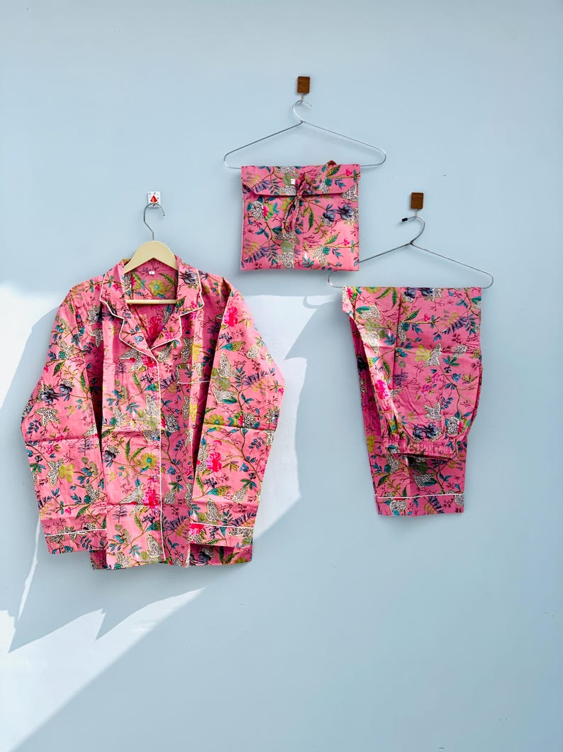Pu&ograve; includere: Set pigiama rosa floreale, composto da una camicia con bottoni, pantaloni e una pochette abbinata. Il set presenta una stampa botanica con accenti verdi, gialli e bianchi. I pigiami sono esposti su grucce.