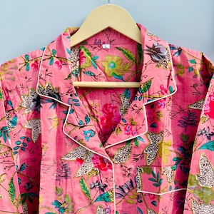 Pu&ograve; includere: Set pigiama rosa floreale con finiture bianche, appeso a una gruccia di legno. Il pigiama presenta un vivace motivo di fiori e foglie nelle tonalit&agrave; del rosa, verde e blu. Il set include una camicia abbottonata con taschino.