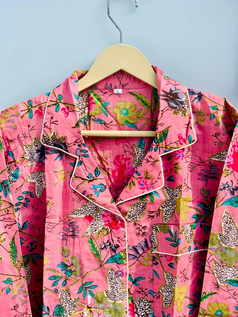 Pu&ograve; includere: Set pigiama rosa floreale con finiture bianche, appeso a una gruccia di legno. Il pigiama presenta un vivace motivo di fiori e foglie nelle tonalit&agrave; del rosa, verde e blu. Il set include una camicia abbottonata con taschino.