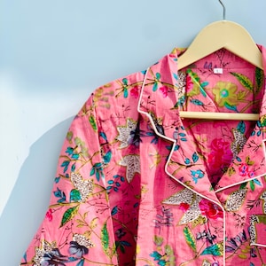 Pu&ograve; includere: Top pigiama rosa a fantasia floreale con bordi bianchi, appeso a una gruccia di legno. Il tessuto presenta un vivace motivo di fiori e foglie nelle tonalit&agrave; del rosa, verde e blu. Il top del pigiama &egrave; abbottonato e ha un colletto.