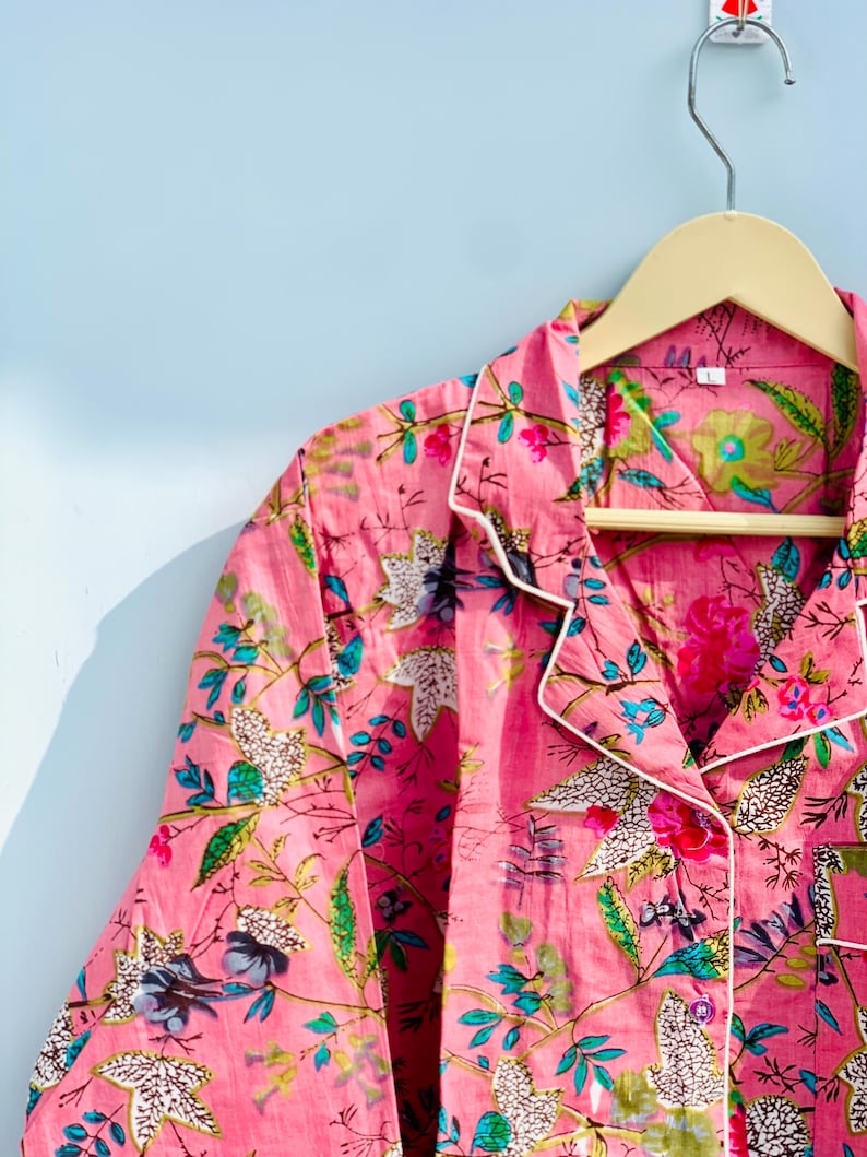 Pu&ograve; includere: Top pigiama rosa a fantasia floreale con bordi bianchi, appeso a una gruccia di legno. Il tessuto presenta un vivace motivo di fiori e foglie nelle tonalit&agrave; del rosa, verde e blu. Il top del pigiama &egrave; abbottonato e ha un colletto.