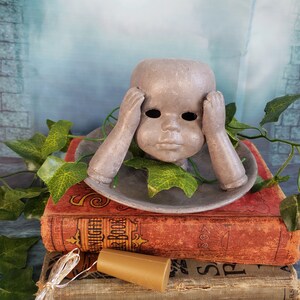 May include: A grey ceramic doll head with hands covering its eyes, sitting on a grey saucer, on top of a stack of vintage books. A string of fairy lights and a brown cork are also in the image.