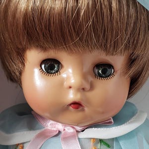 May include: A vintage porcelain doll with brown hair, green eyes, and a light blue dress with pink ribbon and floral embroidery.