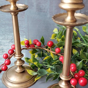 May include: Two gold metal candle holders with a decorative beaded design. The candle holders are on a gray surface with a red berry garland.