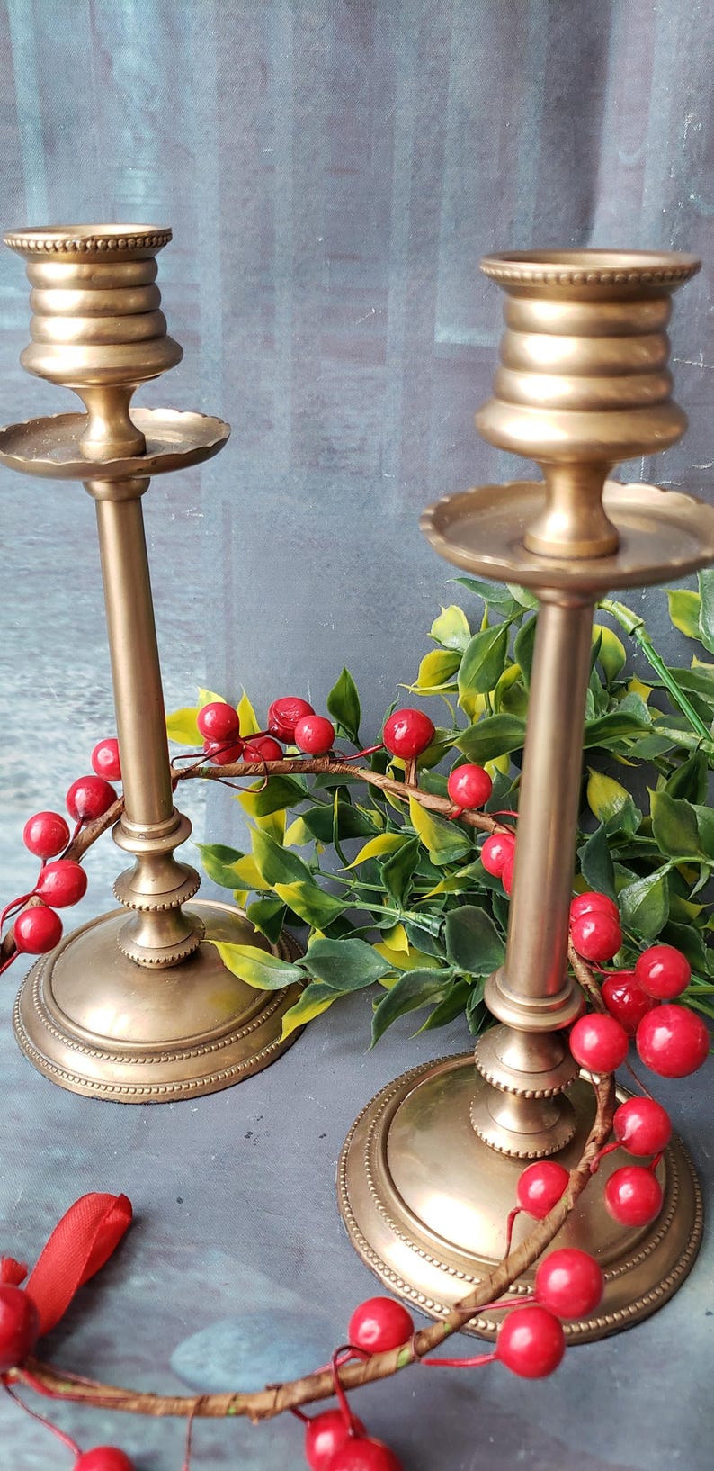May include: Two gold metal candle holders with a decorative beaded design. The candle holders are on a gray surface with a red berry garland.