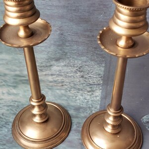 May include: Two gold metal candle holders with a fluted design and a beaded rim. The candle holders are on a gray surface.