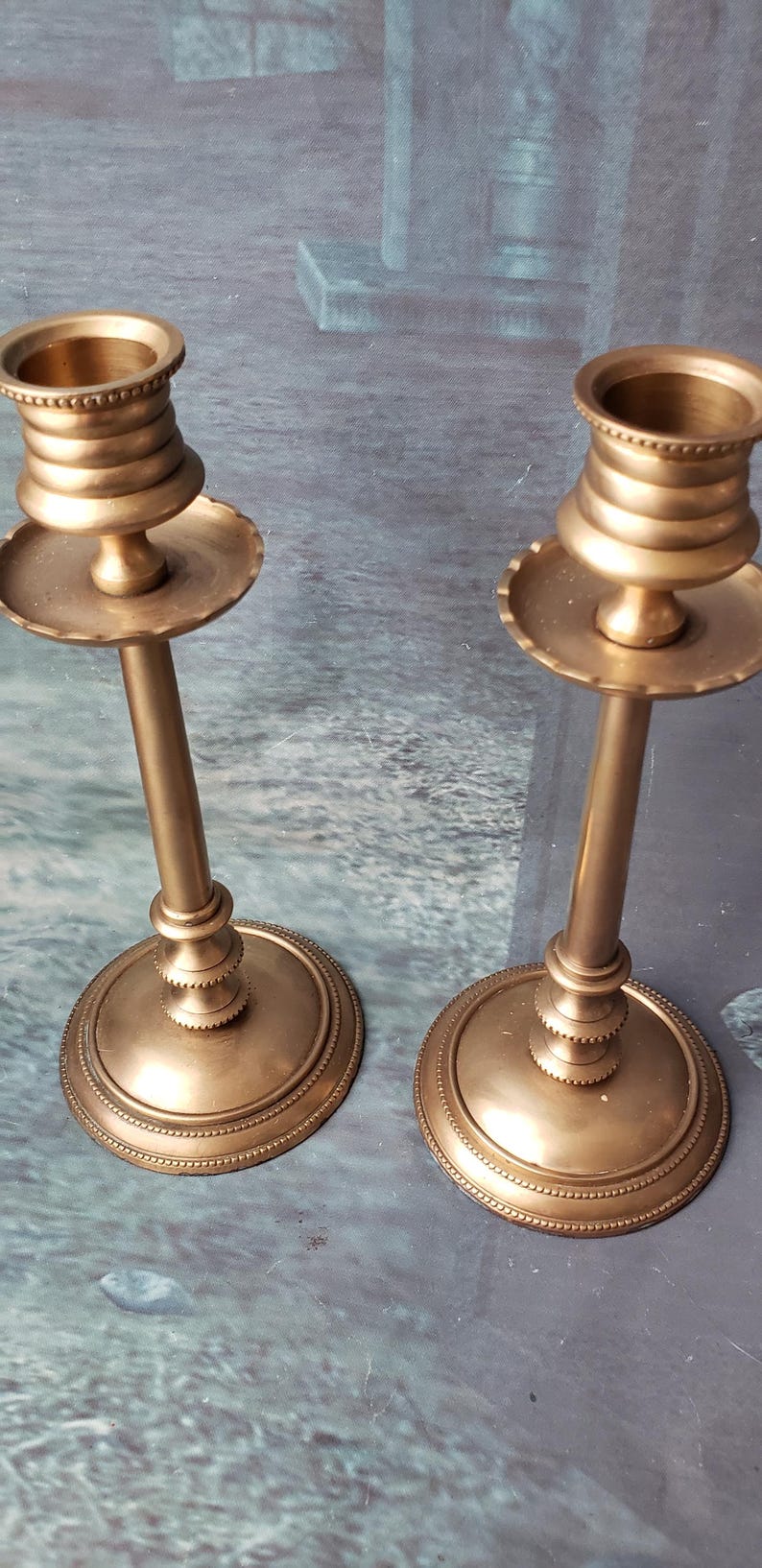 May include: Two gold metal candle holders with a fluted design and a beaded rim. The candle holders are on a gray surface.