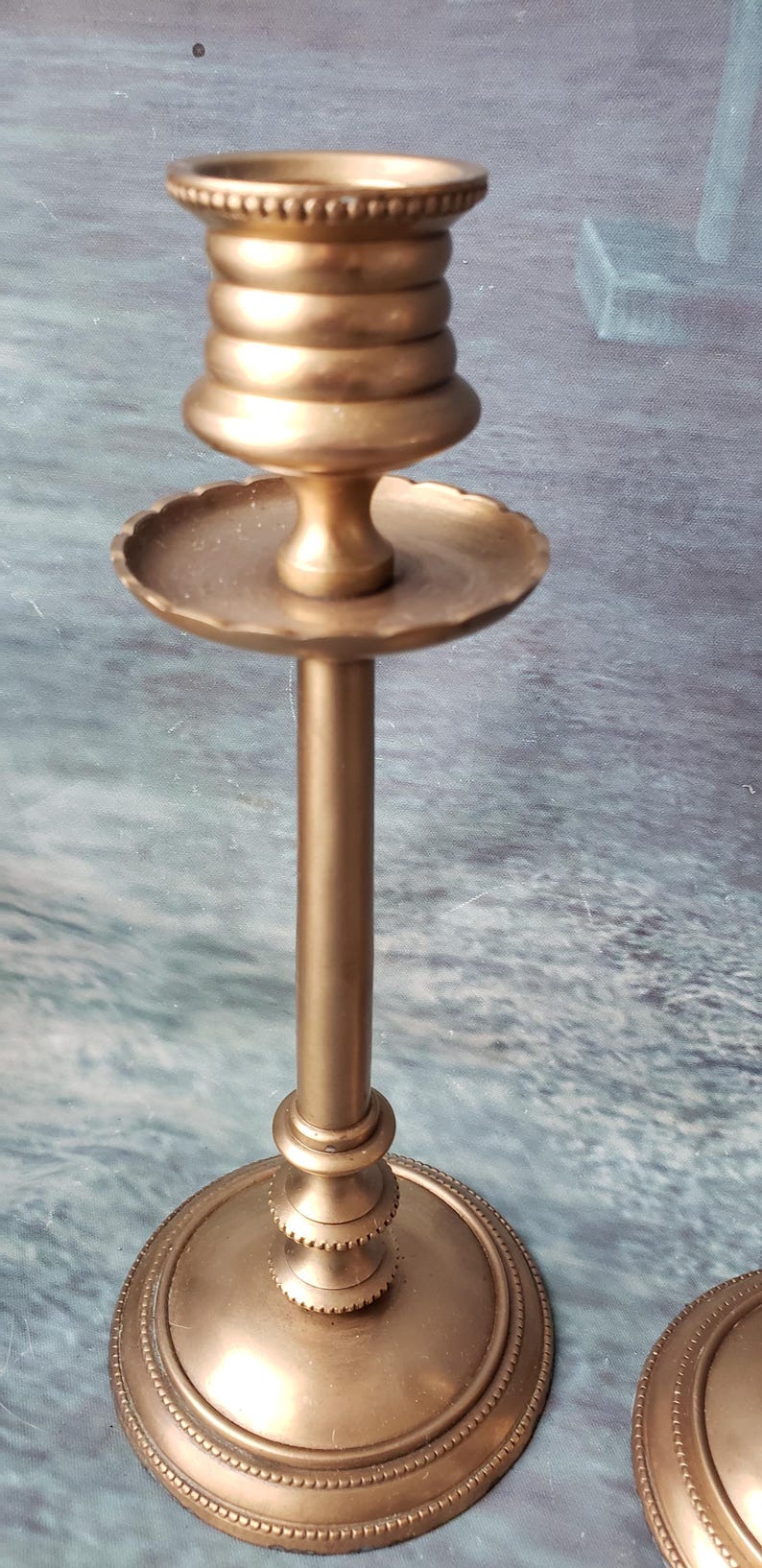 May include: A pair of gold metal candle holders with a decorative design. The candle holders have a round base and a tall, slender stem. The top of the candle holder is a round, open cup.