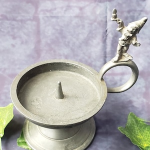 May include: A pewter candle holder featuring a small gnome figure on the handle. The holder has a round base and a circular candle cup with a central spike.