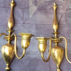 May include: Two antique brass wall sconces. Each sconce has a central body with a decorative top and three candle holders. The sconces have curved arms and are mounted on a wall. The brass has a warm, golden hue.