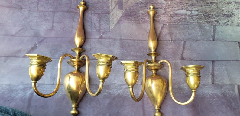 May include: Two antique brass wall sconces. Each sconce has a central body with a decorative top and three candle holders. The sconces have curved arms and are mounted on a wall. The brass has a warm, golden hue.