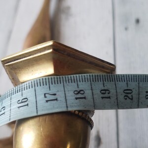 May include: A vintage brass candlestick holder with a square top and a bulbous base. The width of the base is approximately 1.5 inches. The candlestick holder is against a white wooden background.