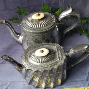 May include: Two antique silver teapots with intricate patterns. The teapots feature ivory-coloured knobs and handles. One teapot is positioned above the other. A measuring tape is in the foreground. The background is a dark, textured surface with green leaves.