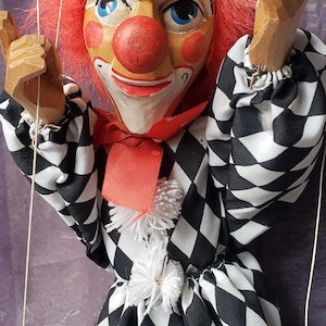 May include: A vintage clown puppet with a painted wooden face, red hair, and a red nose. The puppet wears a black and white diamond-patterned outfit with a red bow tie and white pom-poms. The puppet's arms are made of wood.