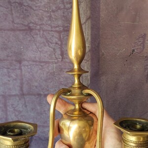 May include: A vintage brass candle holder with two arms and a central stem. The candle holders have a detailed design and are held by curved arms. The brass has a warm, aged patina, suggesting it is an antique.