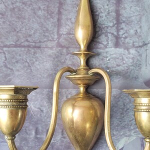 May include: A vintage brass wall sconce with two curved arms and candle holders. The sconce has a central decorative element and a warm, golden hue. The sconce is mounted on a textured, purple-gray wall.