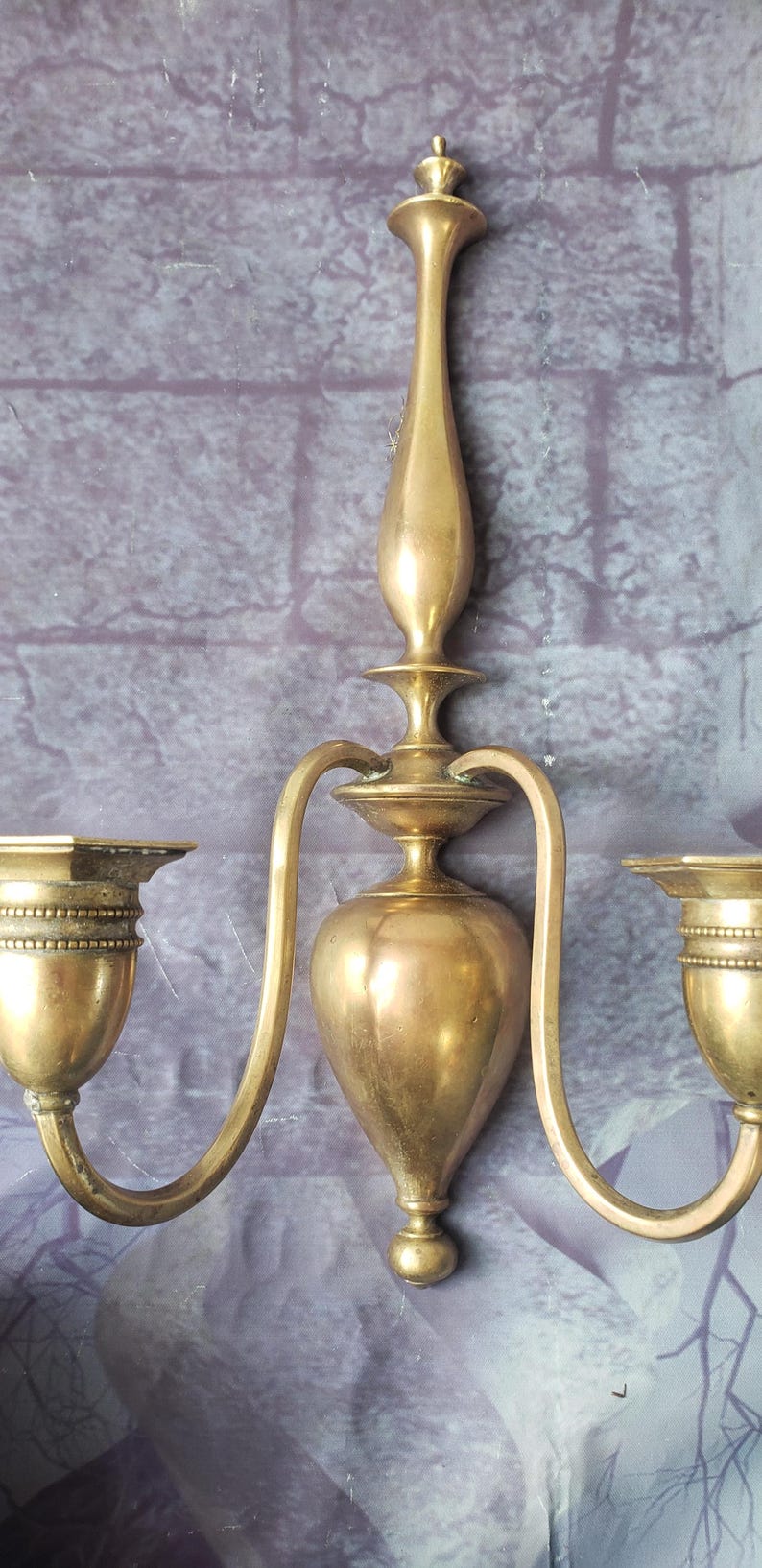 May include: A vintage brass wall sconce with two curved arms and candle holders. The sconce has a central decorative element and a warm, golden hue. The sconce is mounted on a textured, purple-gray wall.