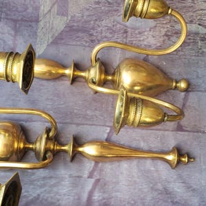 May include: Four antique brass candle holders. Each holder has a curved arm with a candle cup and a decorative base. The brass has a warm, golden hue. The candle holders are arranged on a purple and gray brick surface.