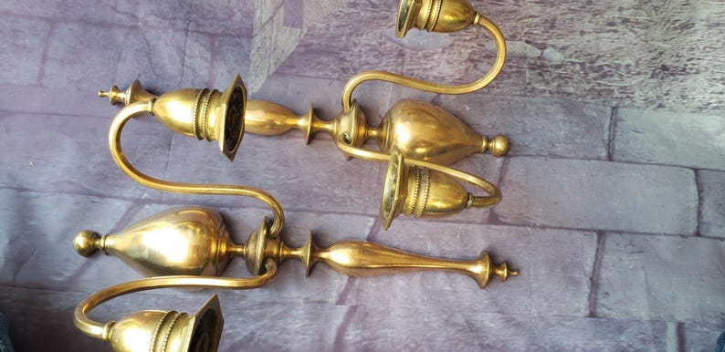 May include: Four antique brass candle holders. Each holder has a curved arm with a candle cup and a decorative base. The brass has a warm, golden hue. The candle holders are arranged on a purple and gray brick surface.