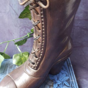 May include: A brown decorative boot with laces, resembling vintage footwear. The boot is made of a dark material and is placed on a blue floral patterned surface. A sprig of greenery is visible.