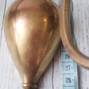 May include: A vintage brass wall sconce with a teardrop-shaped body and curved handle. The sconce is approximately 12 inches tall, with a measurement tape alongside it. The brass has a warm, aged patina.