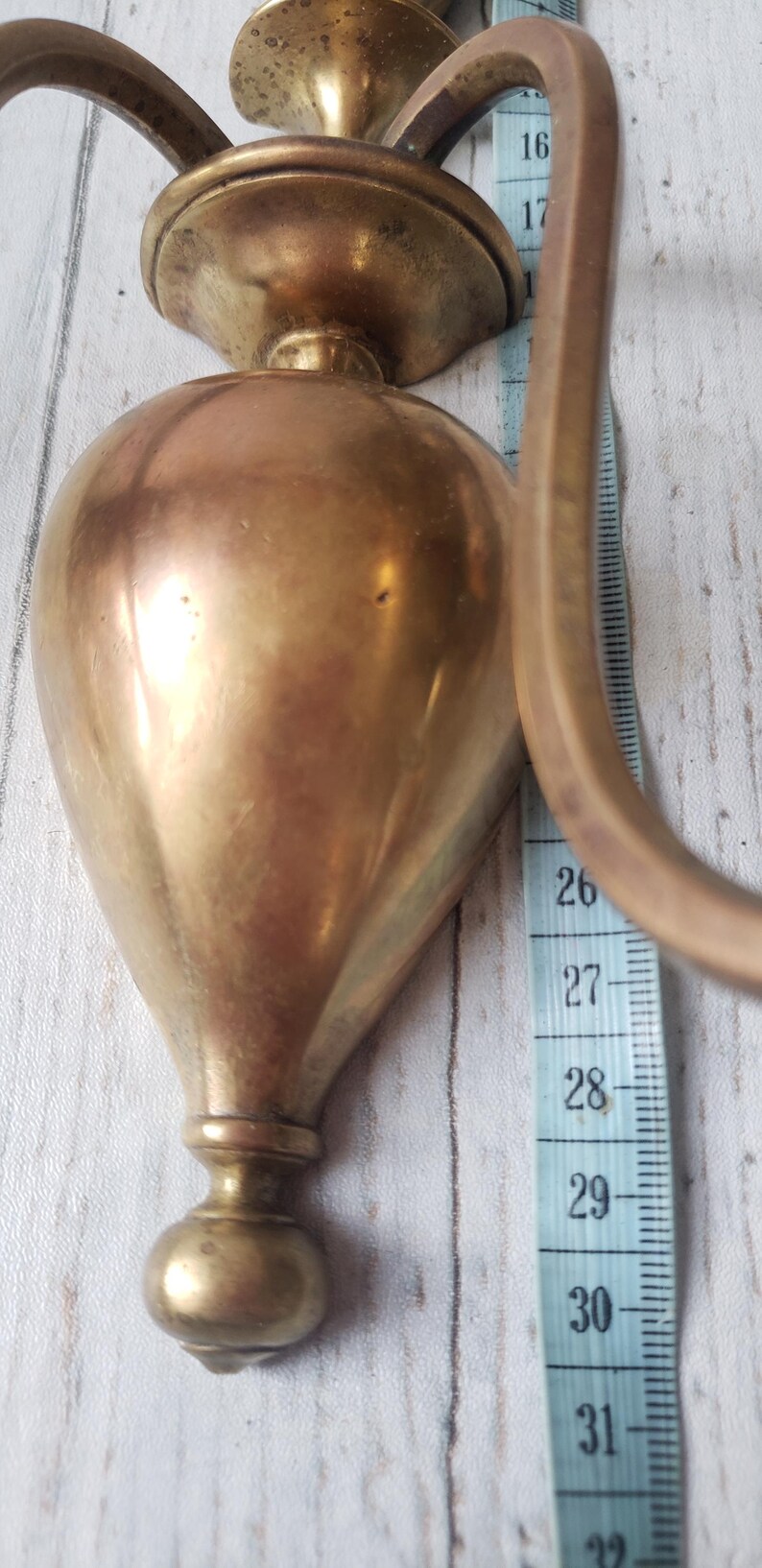 May include: A vintage brass wall sconce with a teardrop-shaped body and curved handle. The sconce is approximately 12 inches tall, with a measurement tape alongside it. The brass has a warm, aged patina.