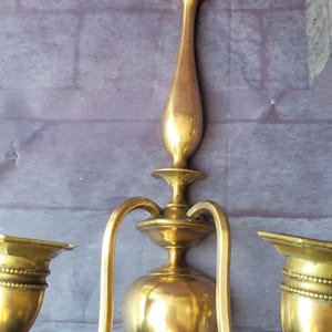 May include: A vintage brass wall sconce with two candle holders. The sconce has a central, elongated body with curved arms extending to the candle cups. The finish is a warm, aged gold color, suggesting an antique piece.