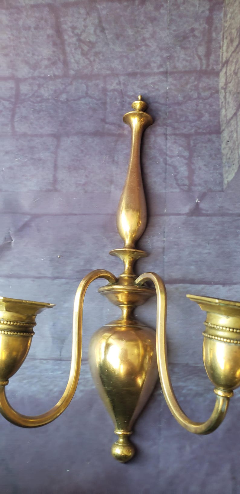 May include: A vintage brass wall sconce with two candle holders. The sconce has a central, elongated body with curved arms extending to the candle cups. The finish is a warm, aged gold color, suggesting an antique piece.